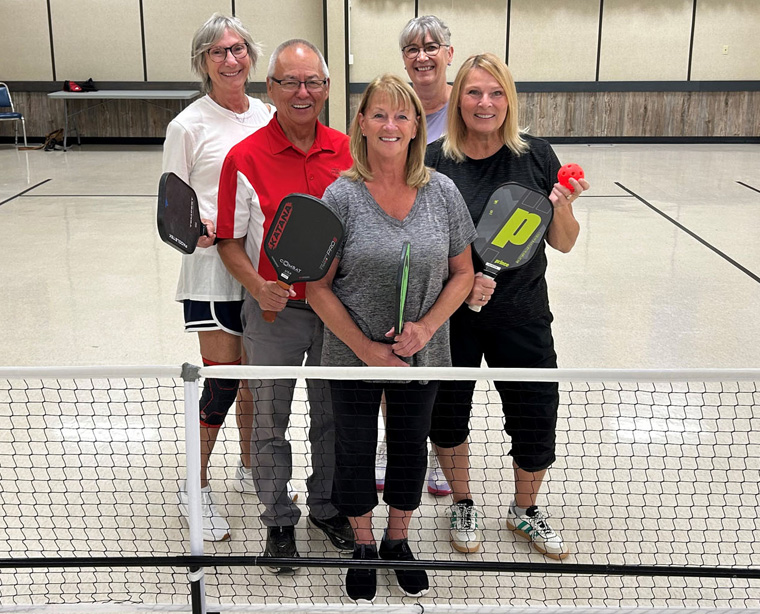 Image of a group of 5 people in sport clothes standing in front of a net ready to play pickleball